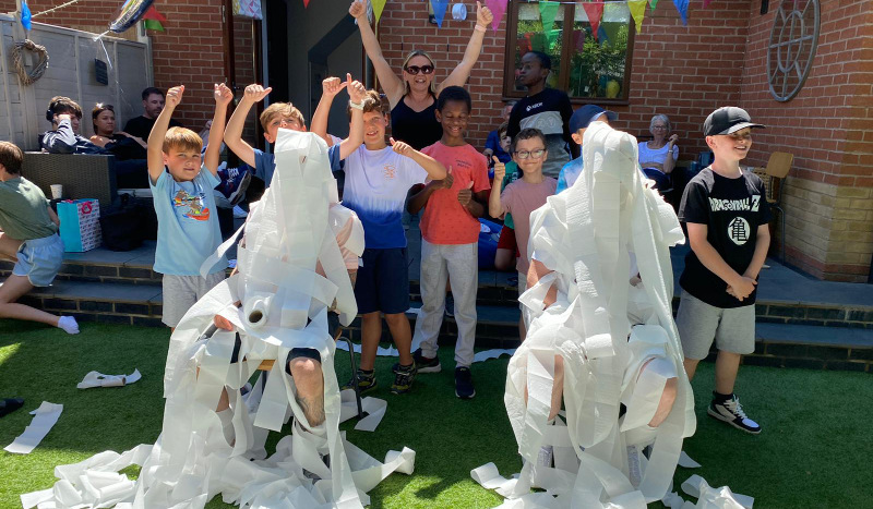 group of children have wrapped two adults sitting on chairs in toilet paper game