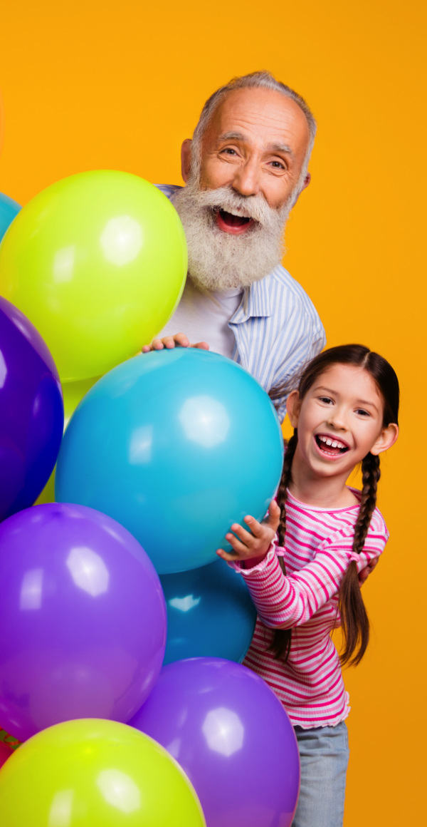 Happy grandfather and his young granddaughters posing together in casual outfits on with colourful balloons